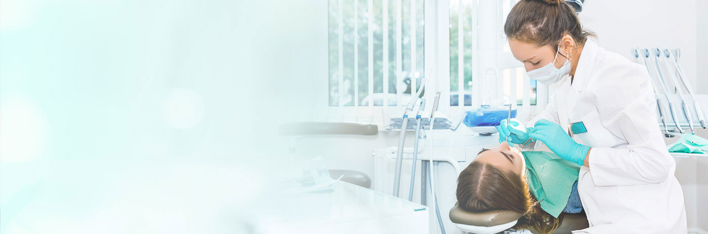 A person with a mask on, likely a dentist, working at a dental station with various equipment around.